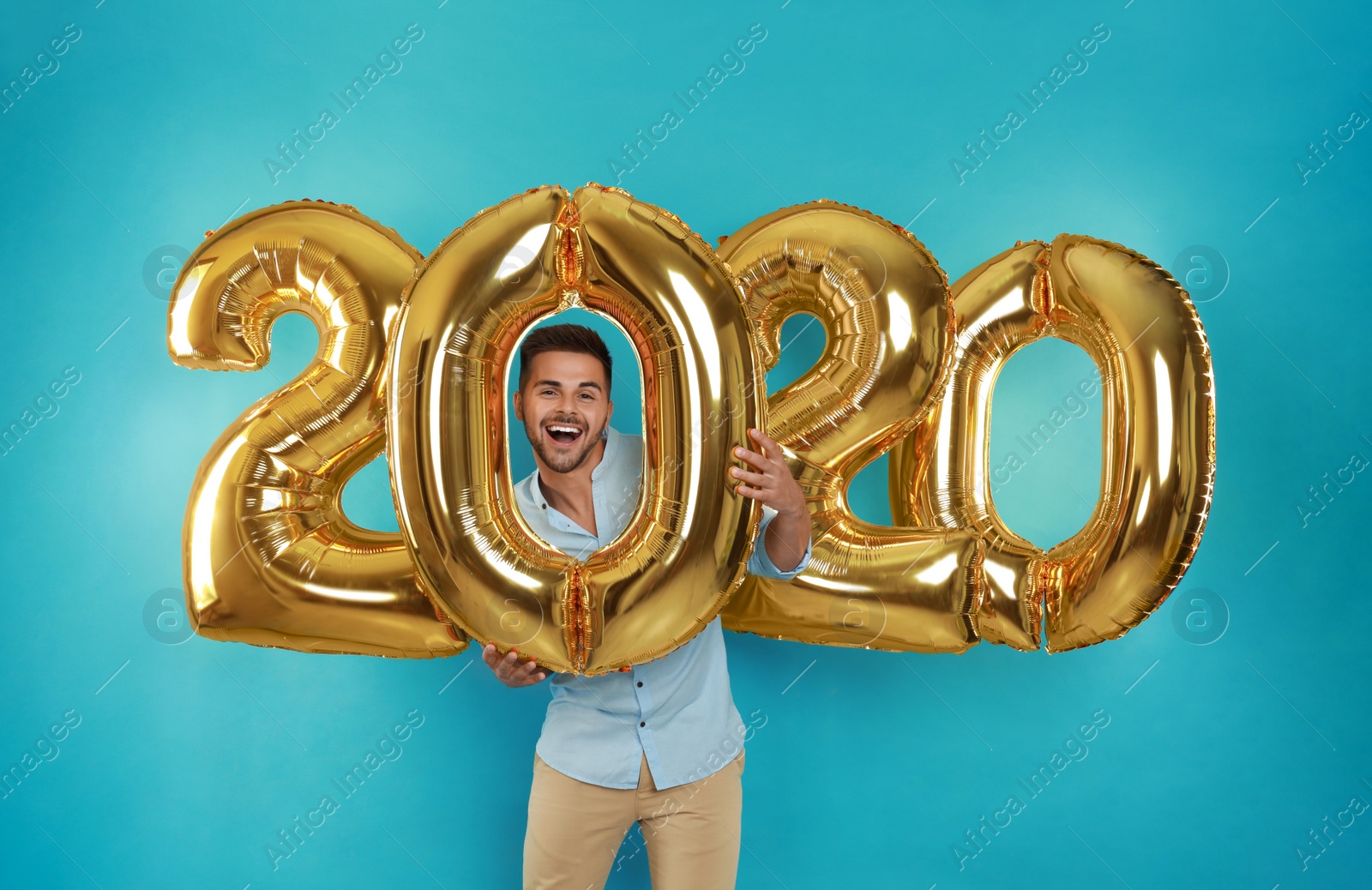 Photo of Happy young man with golden 2020 balloons on turquoise background. New Year celebration