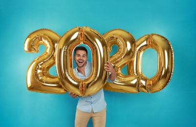 Photo of Happy young man with golden 2020 balloons on turquoise background. New Year celebration