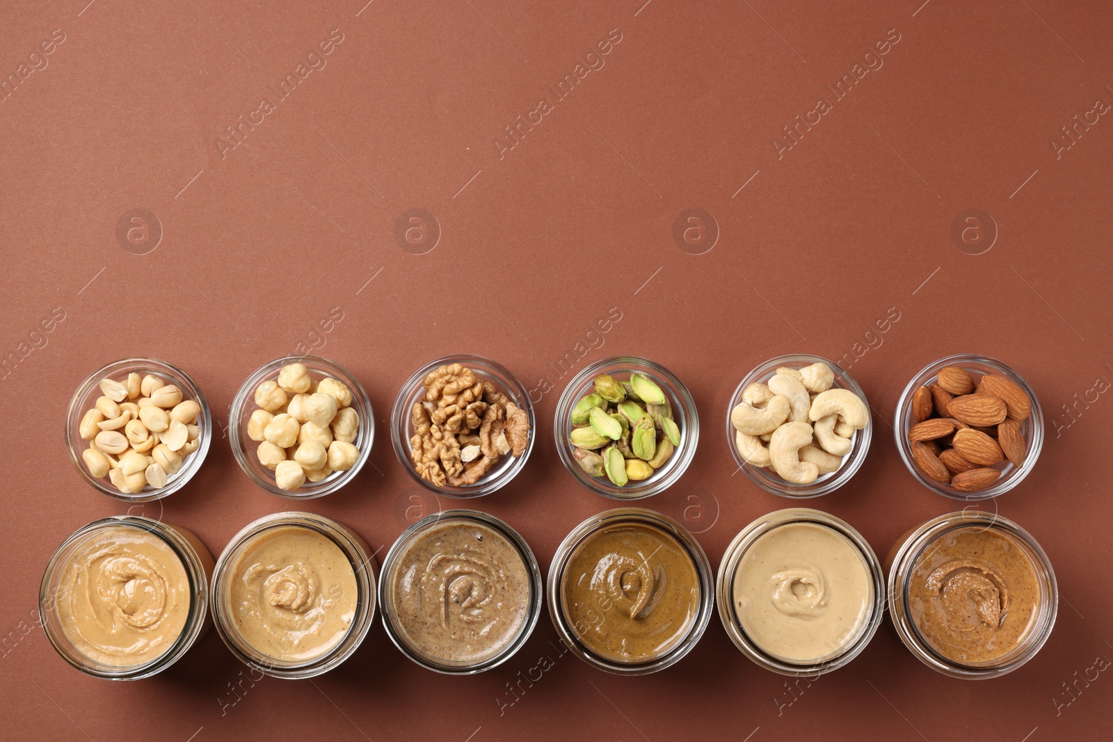 Photo of Many tasty nut butters in jars and nuts on brown table, flat lay. Space for text