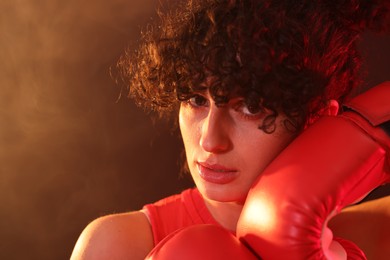 Photo of Beautiful young woman with boxing gloves on color background in neon lights and smoke, closeup. Space for text