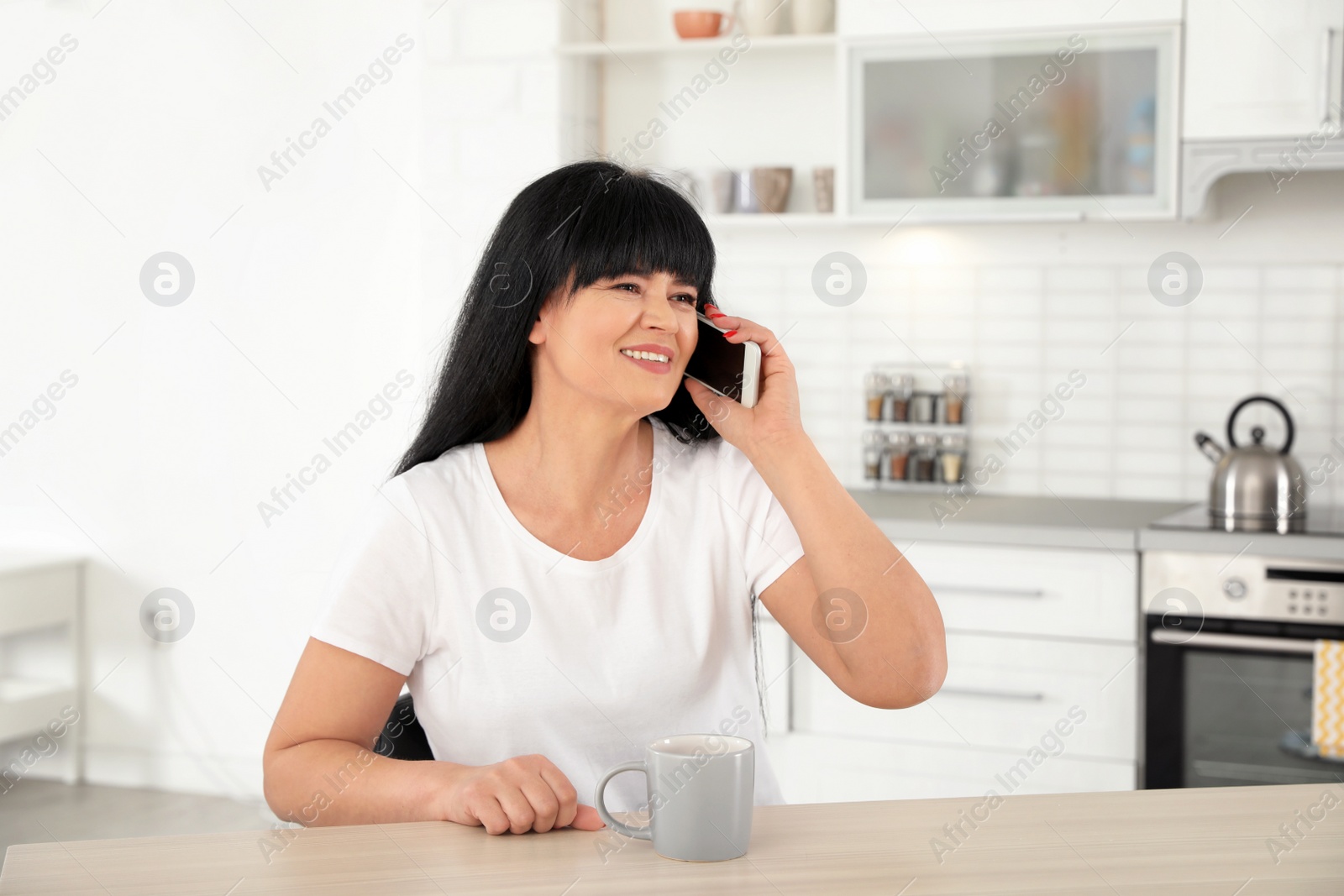 Photo of Mature woman with cup of drink and smartphone sitting at table indoors