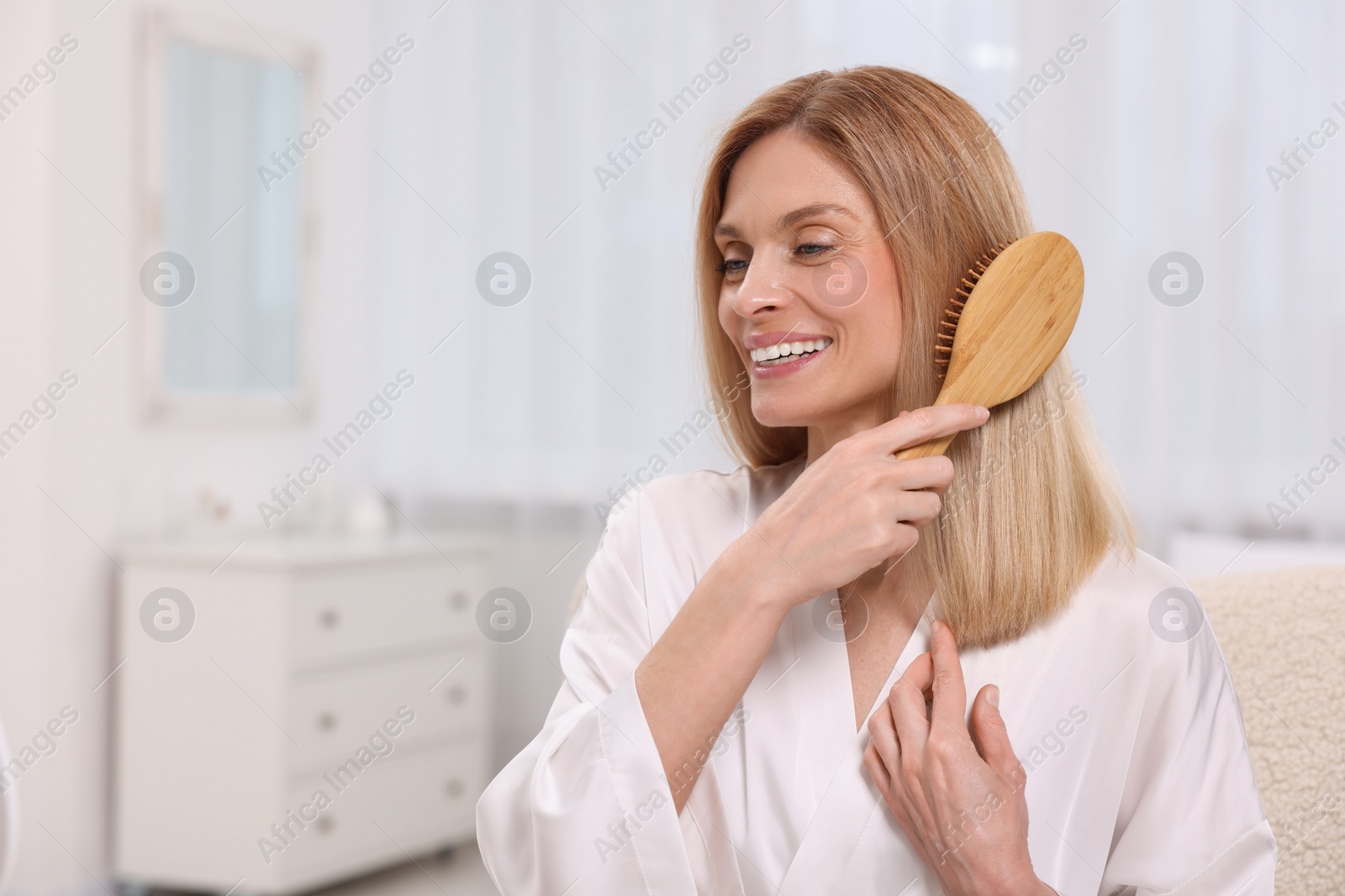 Photo of Beautiful happy woman brushing her hair in bedroom, space for text