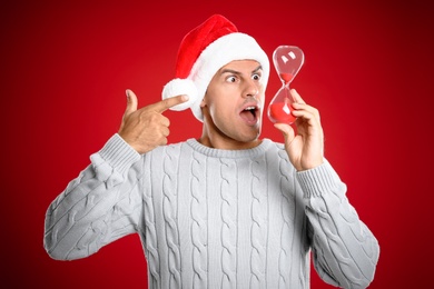 Photo of Man in Santa hat with hourglass on red background. Christmas countdown