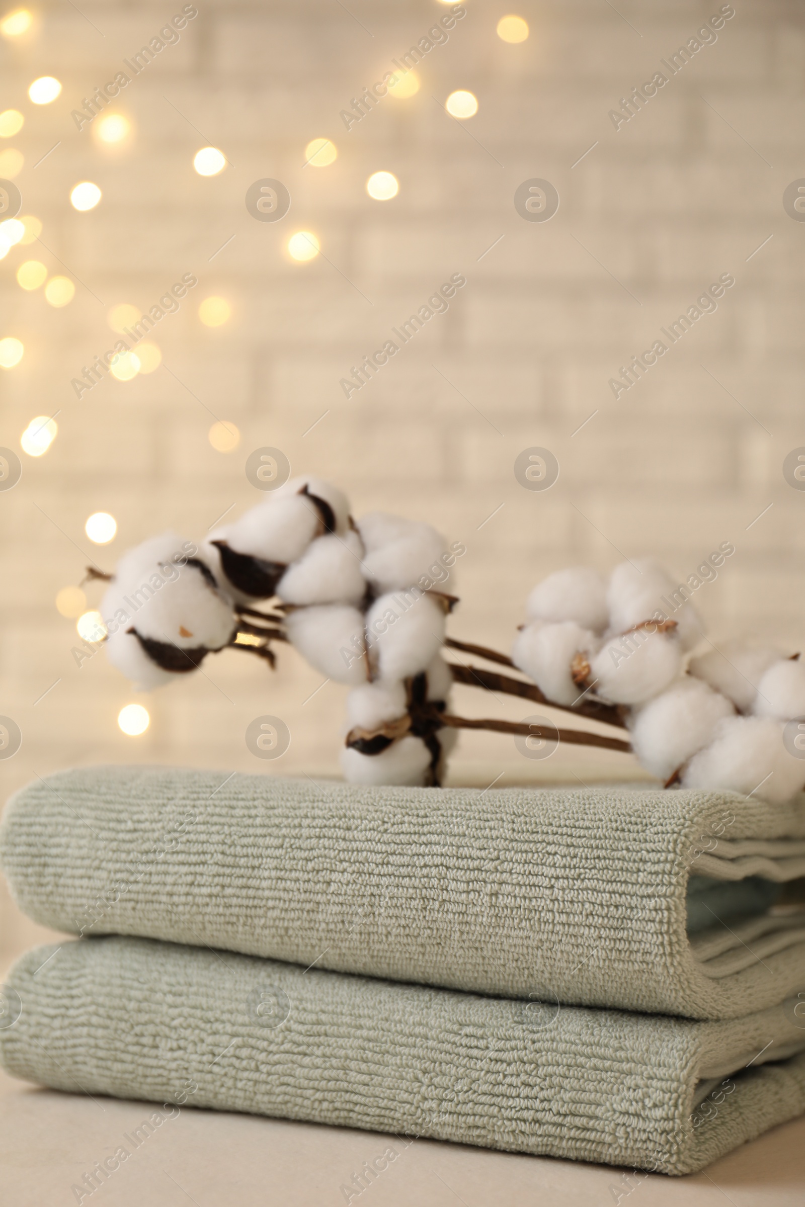 Photo of Stacked soft towels and cotton branches on white table near brick wall indoors, closeup. Space for text