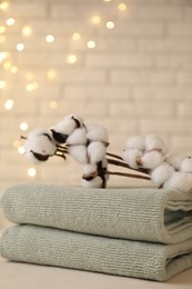 Photo of Stacked soft towels and cotton branches on white table near brick wall indoors, closeup. Space for text
