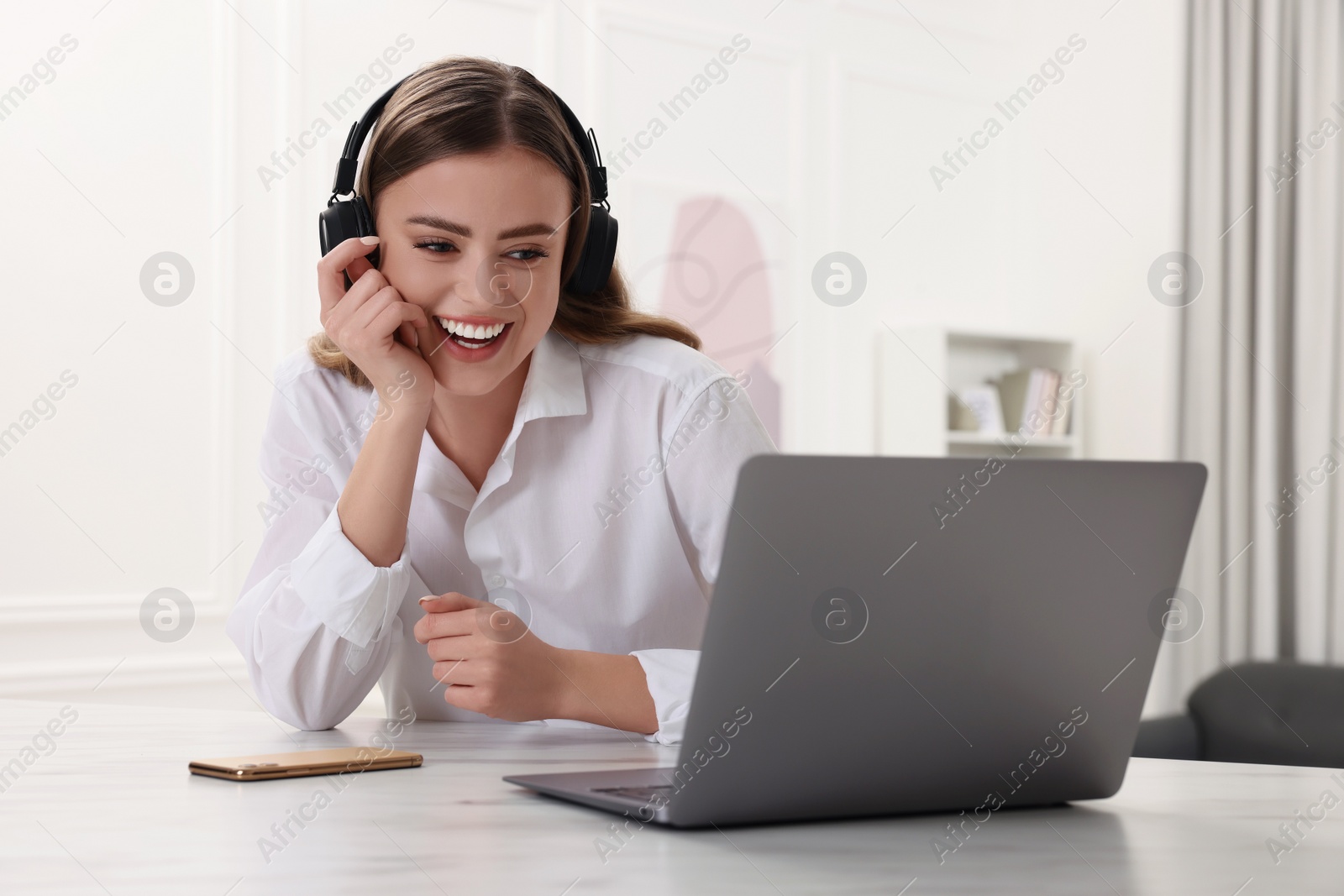Photo of Happy woman with headphones using laptop at white table in room