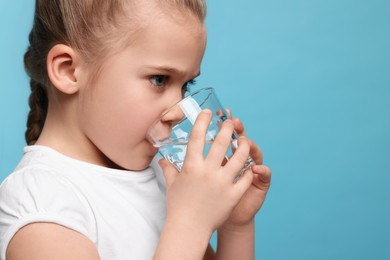 Cute little girl drinking fresh water from glass on light blue background, space for text