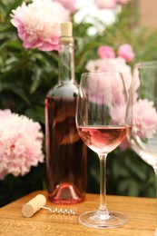 Glasses and bottle with rose wine on wooden table near beautiful peonies