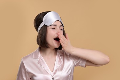 Tired young woman with sleep mask yawning on beige background. Insomnia problem