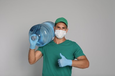 Photo of Courier in medical mask with bottle for water cooler showing thumb up on light grey background. Delivery during coronavirus quarantine