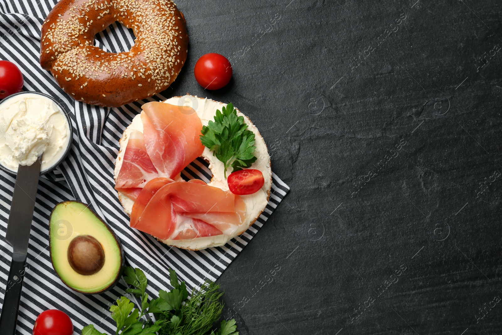 Photo of Delicious bagel with cream cheese, jamon, tomato and parsley on black table, flat lay. Space for text