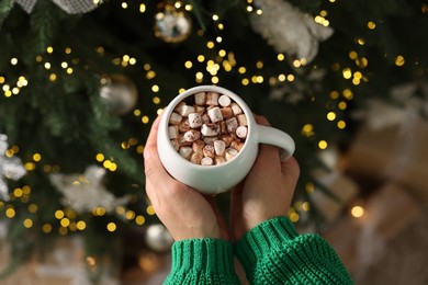 Woman holding cup of delicious Christmas cocoa with marshmallows near fir tree, above view
