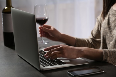 Photo of Woman with glass of wine using laptop at table indoors, closeup. Loneliness concept