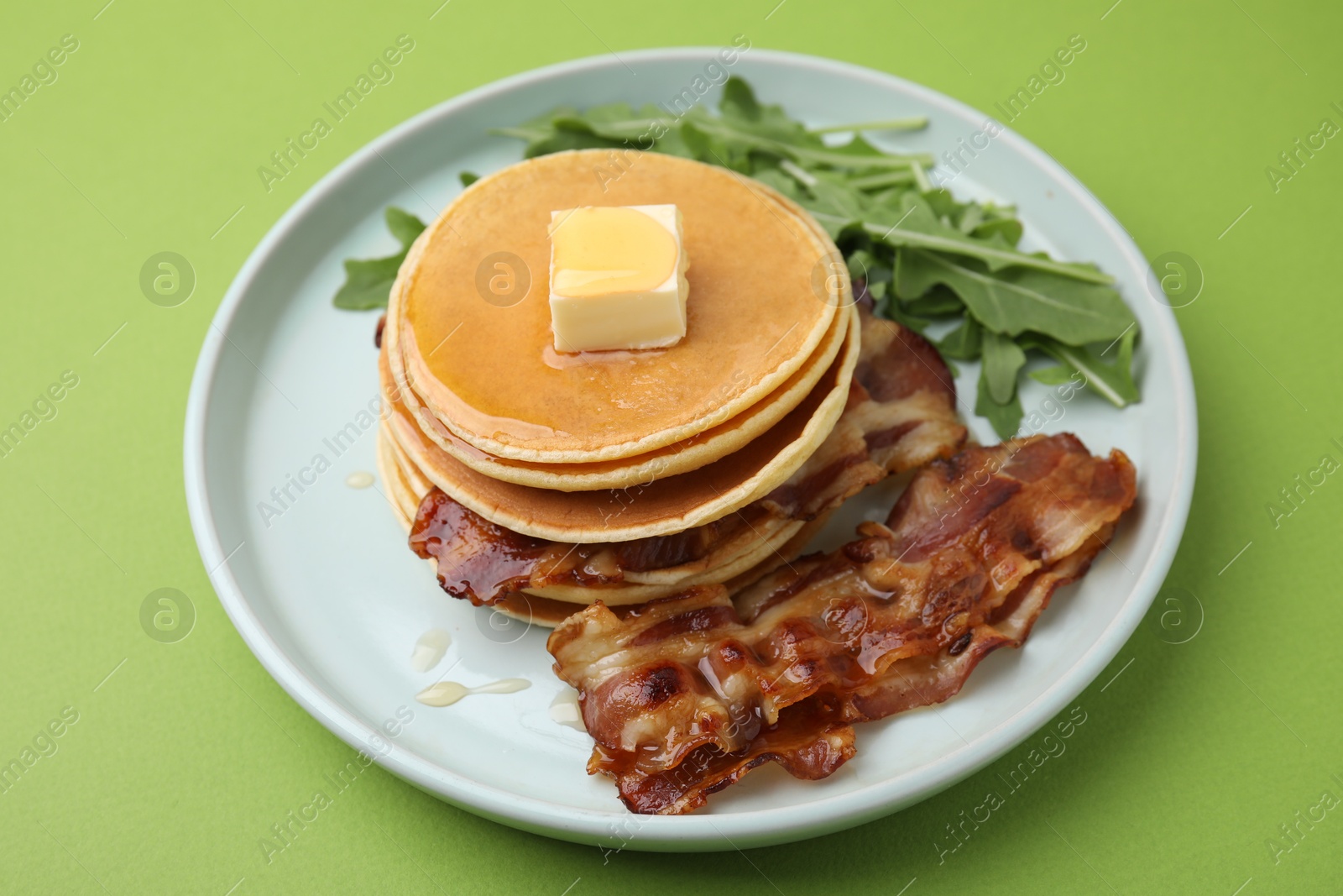 Photo of Tasty pancakes with butter, fried bacon and fresh arugula on light green background