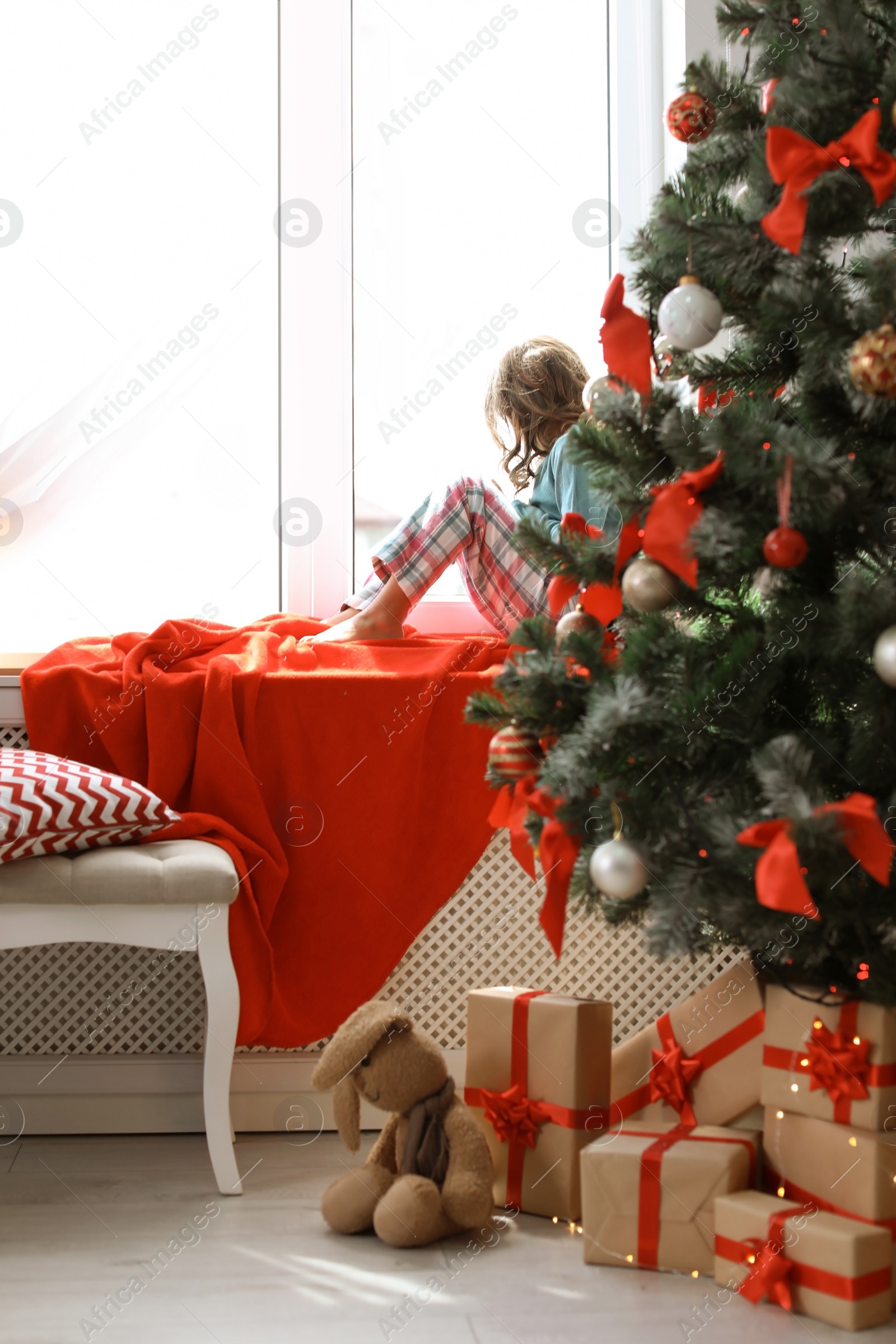 Photo of Cute little child sitting on windowsill near Christmas tree at home