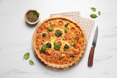 Delicious homemade vegetable quiche, basil leaves, seasoning and knife on white marble table, flat lay