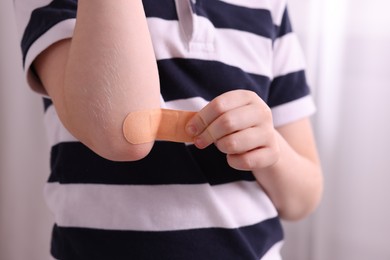Photo of Little boy putting sticking plaster onto elbow indoors, closeup