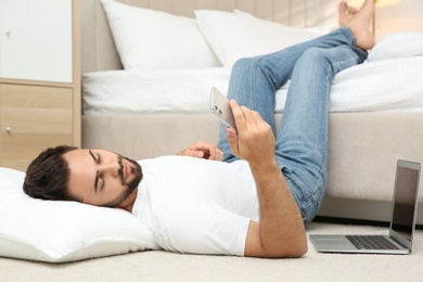 Photo of Lazy young man with laptop and smartphone lying on floor at home