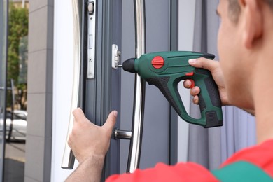 Photo of Handyman with screw gun repairing door lock outdoors, closeup