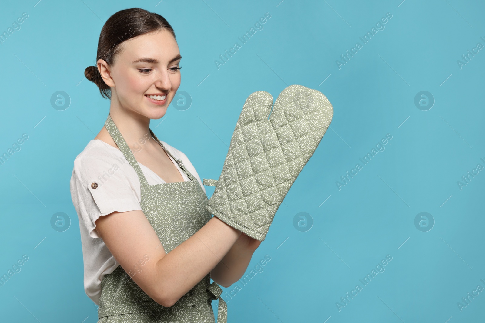 Photo of Beautiful young woman in clean apron with pattern and oven glove on light blue background. Space for text