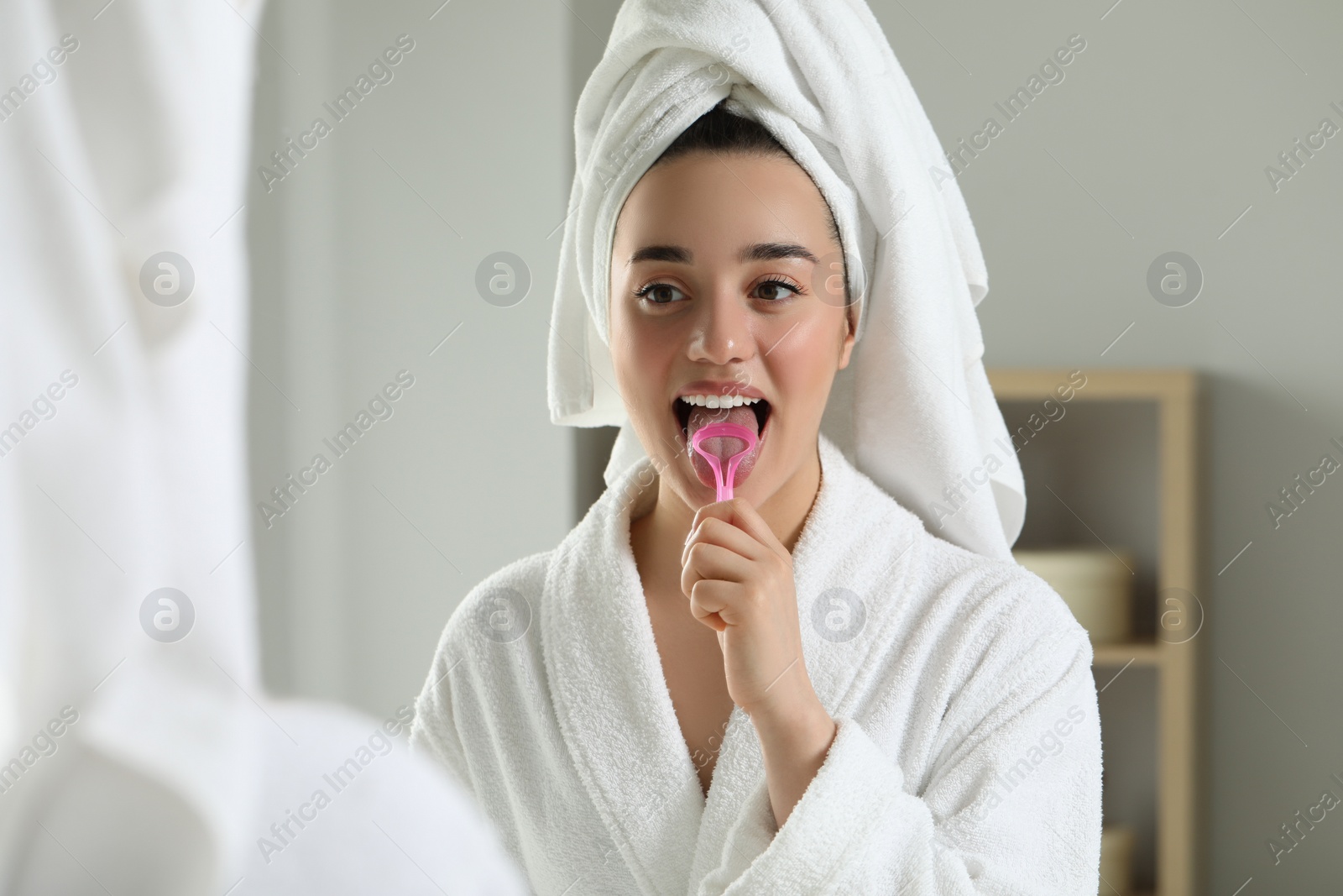 Photo of Happy woman brushing her tongue with cleaner near mirror in bathroom