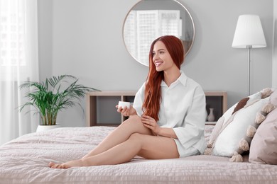 Photo of Beautiful young woman applying body cream onto legs in bedroom, space for text