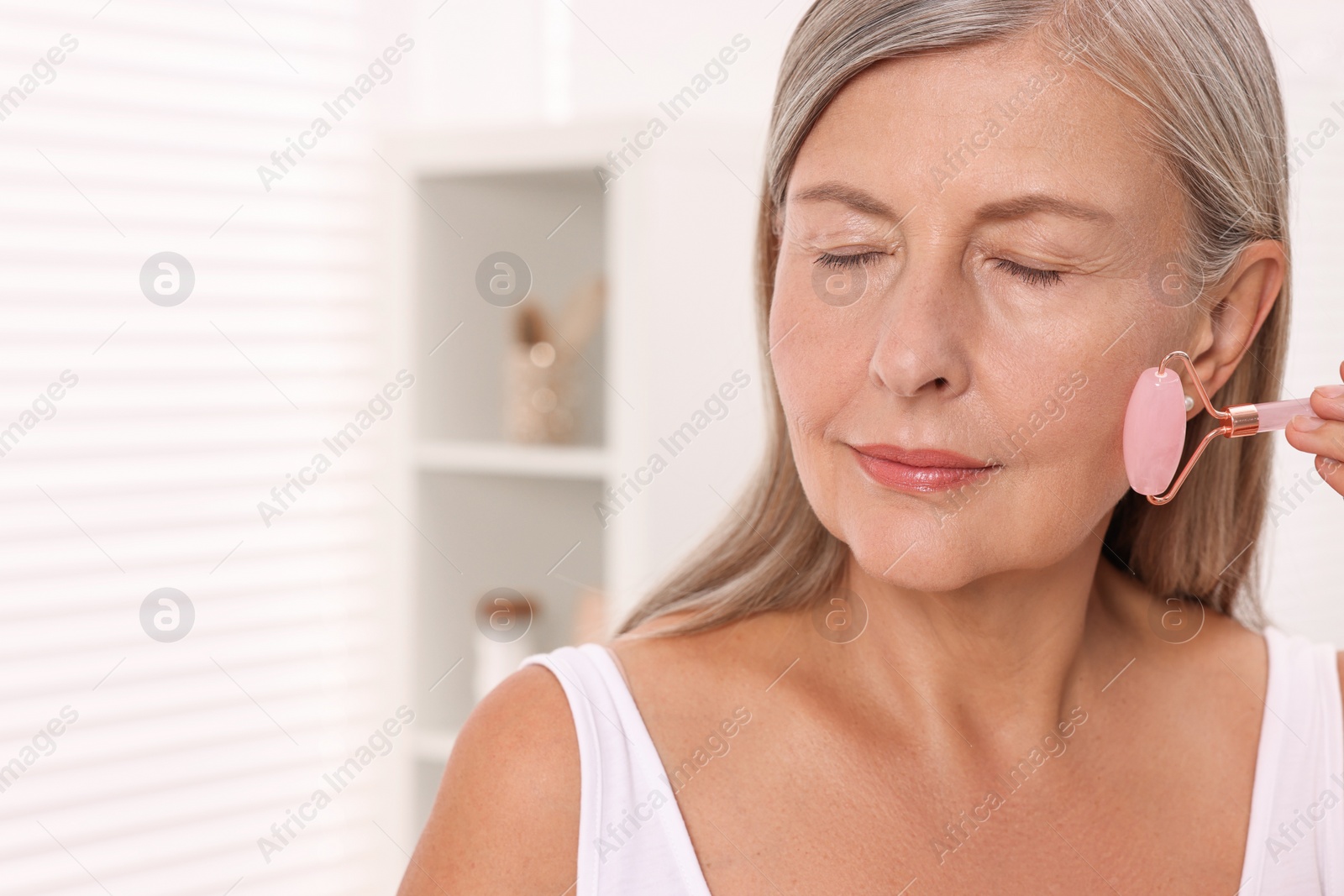 Photo of Woman massaging her face with rose quartz roller in bathroom. Space for text