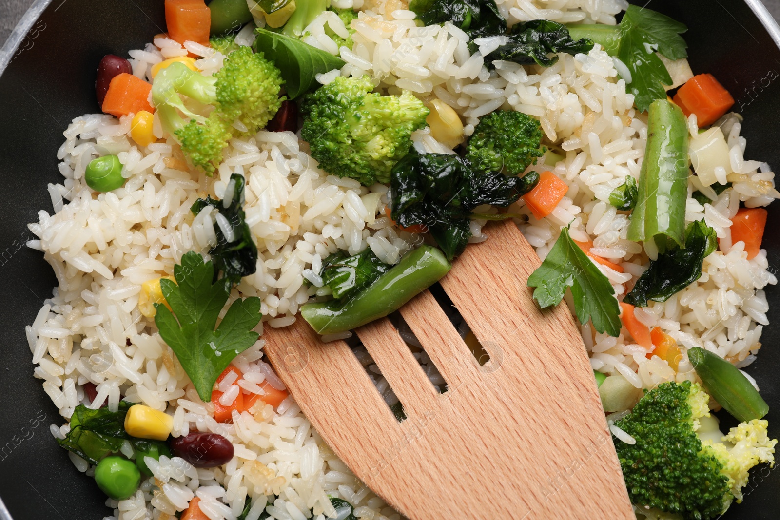 Photo of Tasty fried rice with vegetables and wooden spatula in frying pan, top view