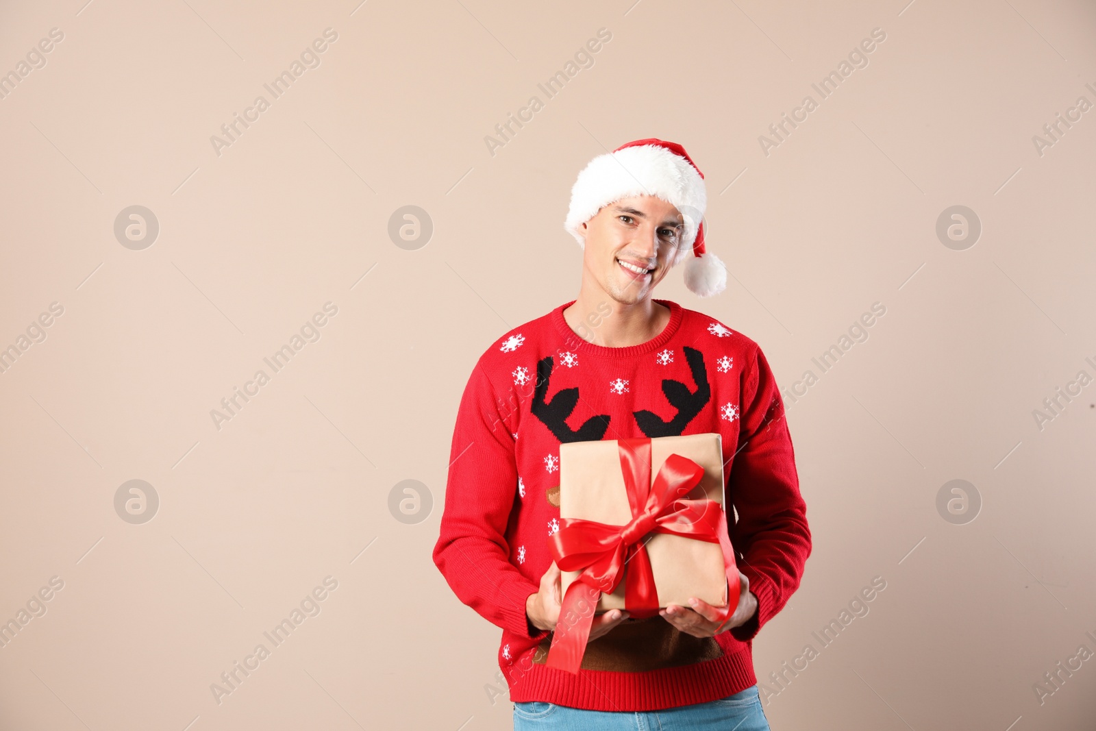 Photo of Happy man in Christmas sweater and Santa hat holding gift box on beige background