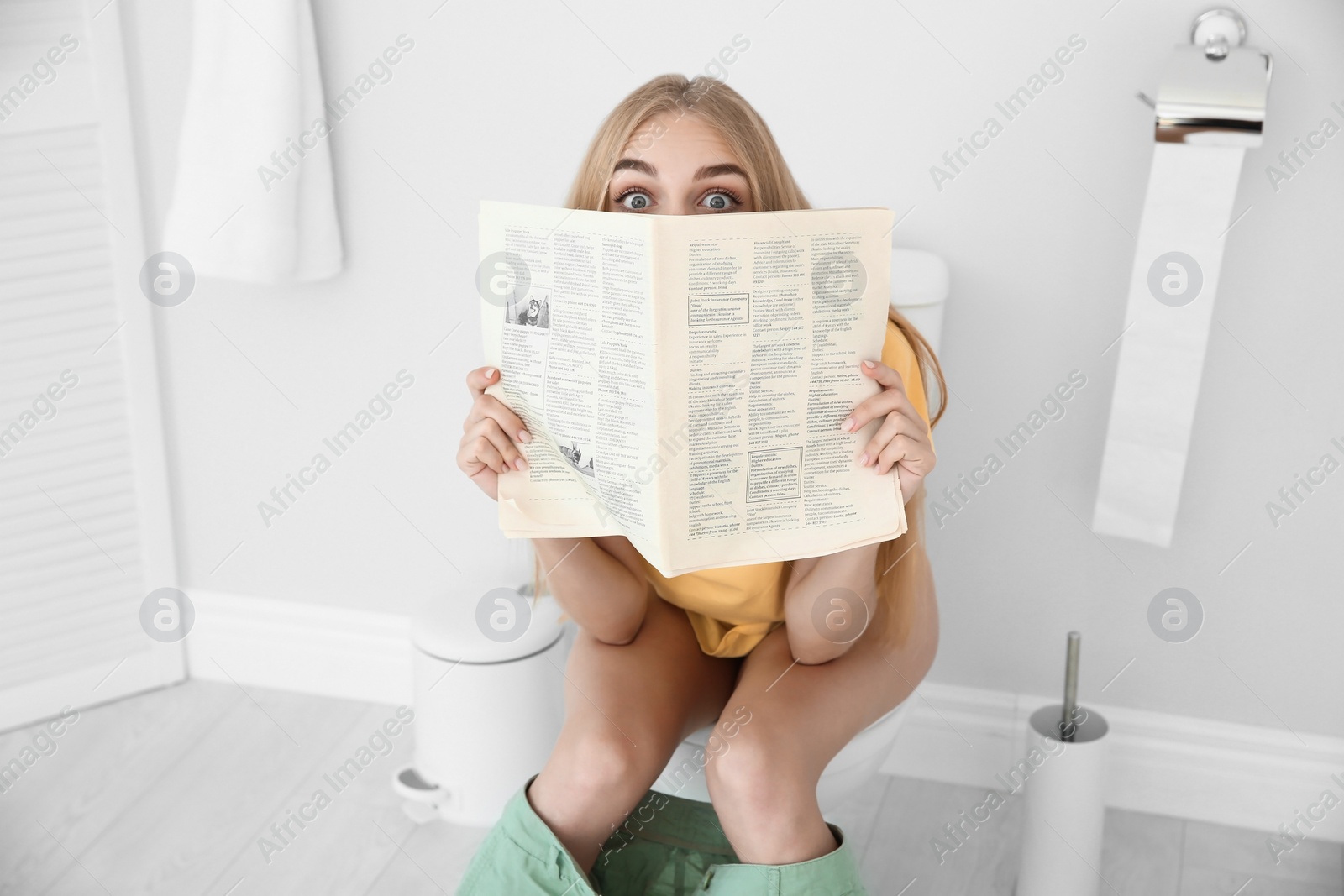 Photo of Young woman reading newspaper while sitting on toilet bowl at home
