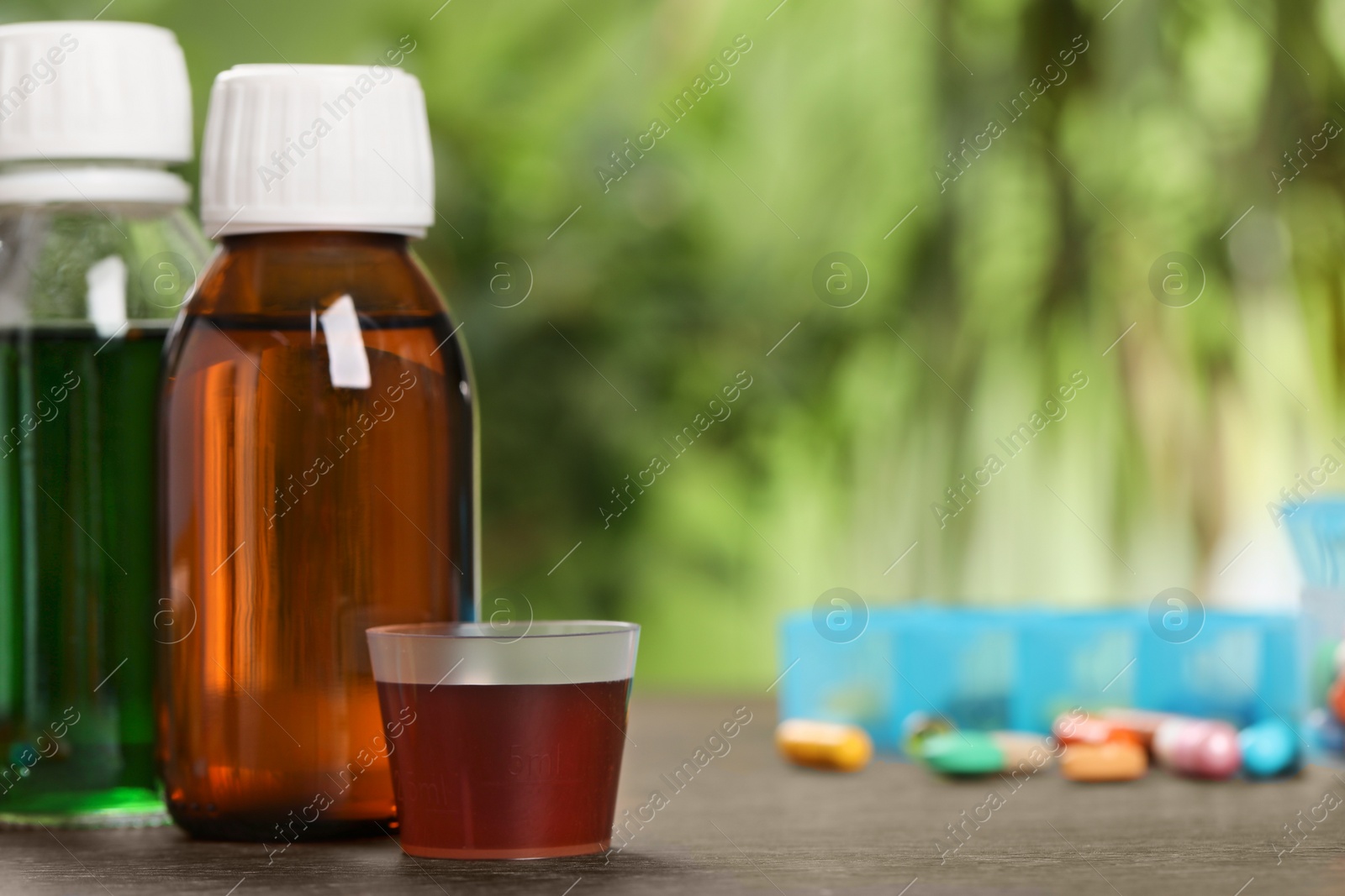 Photo of Bottles of syrup and measuring cup on wooden table, space for text. Cold medicine