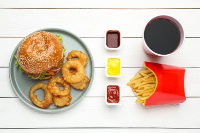 Tasty burger, fried onion rings, French fries, sauces and refreshing drink on white wooden table, flat lay. Fast food