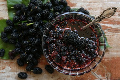 Delicious ripe black mulberries and bowl of sweet jam on wooden table, flat lay