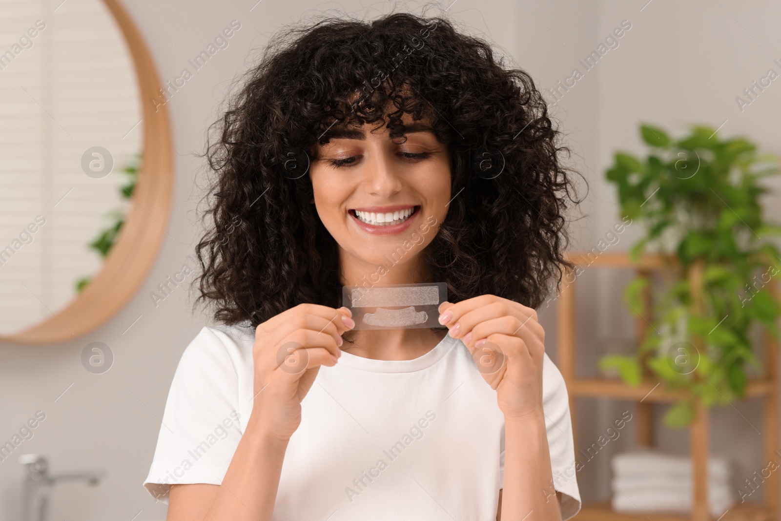 Photo of Young woman holding teeth whitening strips in bathroom