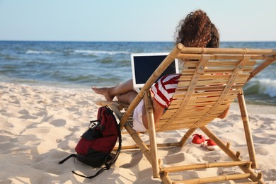 Photo of African American woman working on laptop in sunbed at beach, back view