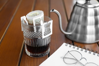 Photo of Glass with drip coffee bag and notebook on wooden table, closeup