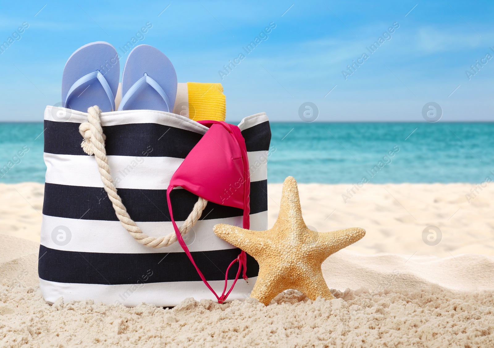 Image of Stylish bag with different accessories on sandy beach near ocean