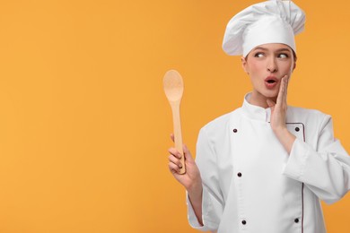 Surprised woman chef in uniform holding wooden spoon on orange background, space for text