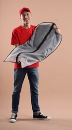 Photo of Dry-cleaning delivery. Happy courier holding garment cover with clothes on beige background