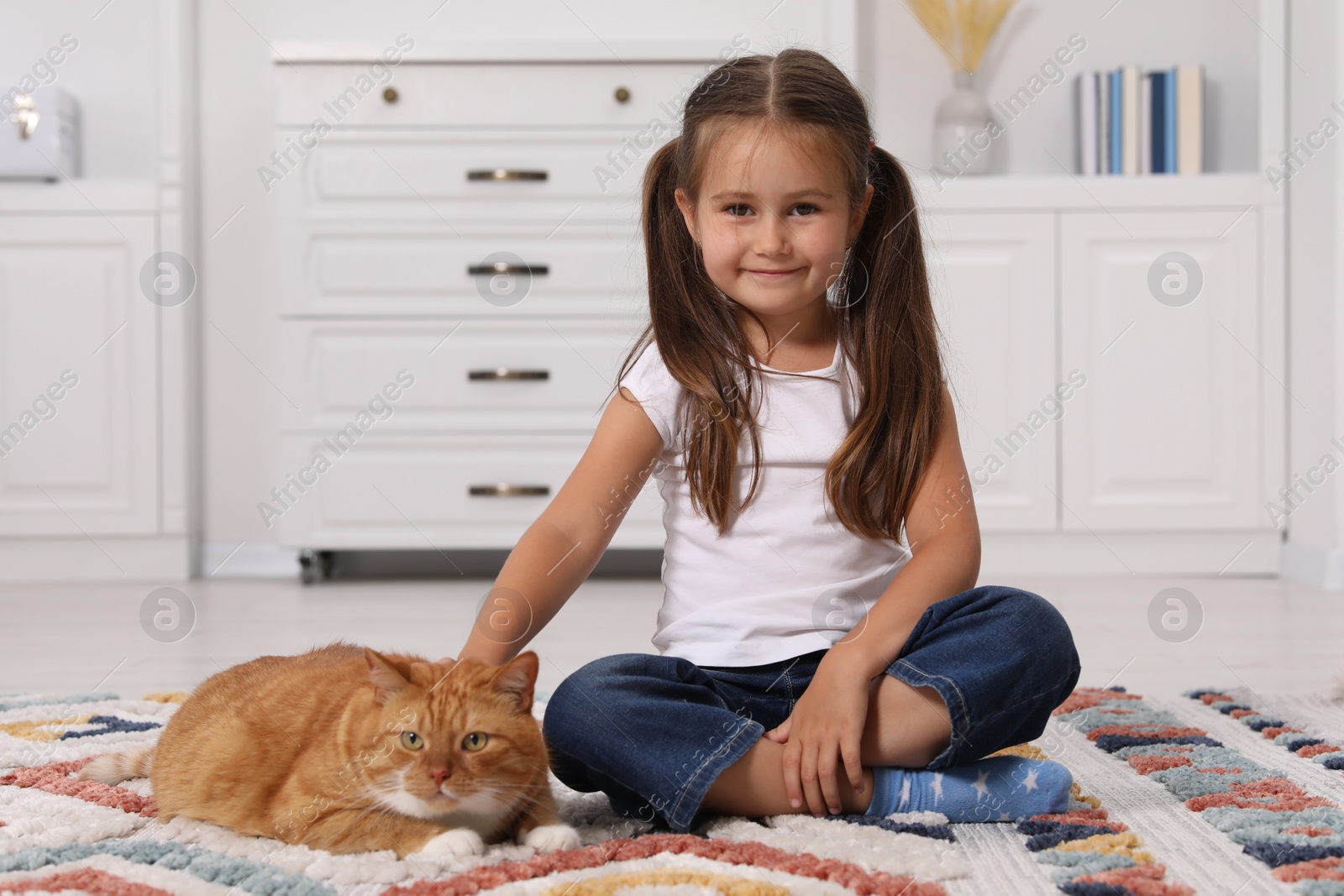 Photo of Smiling little girl petting cute ginger cat on carpet at home