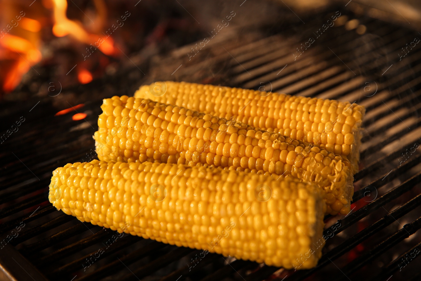 Photo of Cooking delicious fresh corn cobs on grilling grate in oven with burning firewood, closeup