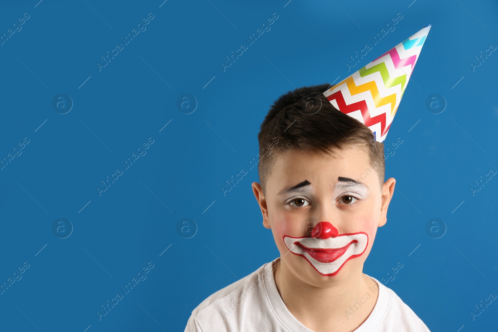 Photo of Preteen boy with clown makeup and party hat on blue background, space for text. April fool's day