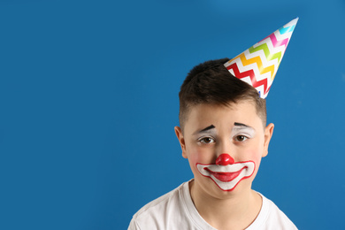 Preteen boy with clown makeup and party hat on blue background, space for text. April fool's day