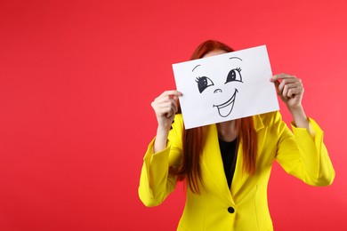 Photo of Woman hiding behind sheet of paper with happy face on red background, space for text