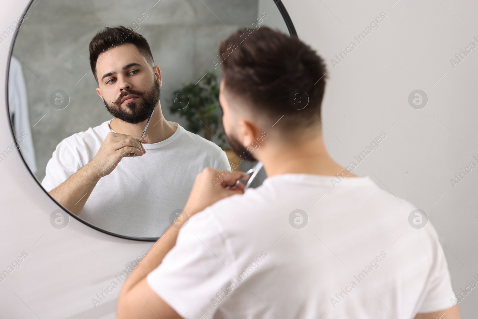 Photo of Handsome young man trimming beard with scissors near mirror in bathroom