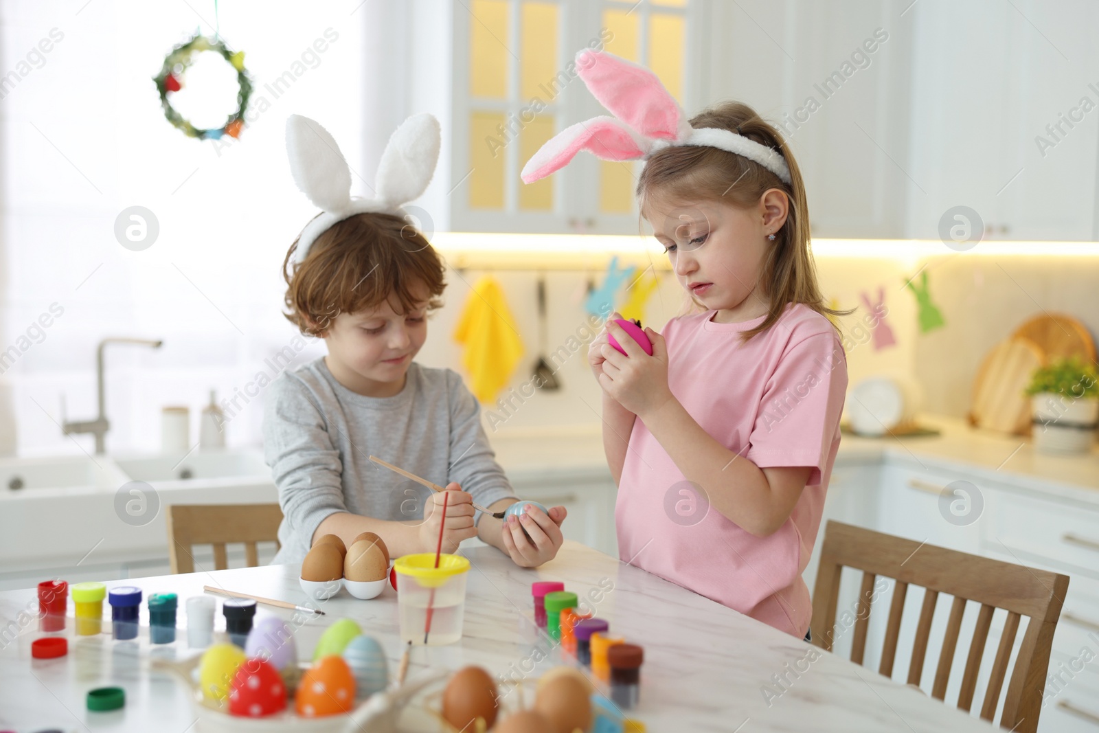 Photo of Easter celebration. Cute children with bunny ears painting eggs at white marble table in kitchen