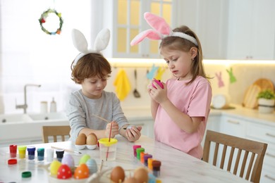 Photo of Easter celebration. Cute children with bunny ears painting eggs at white marble table in kitchen