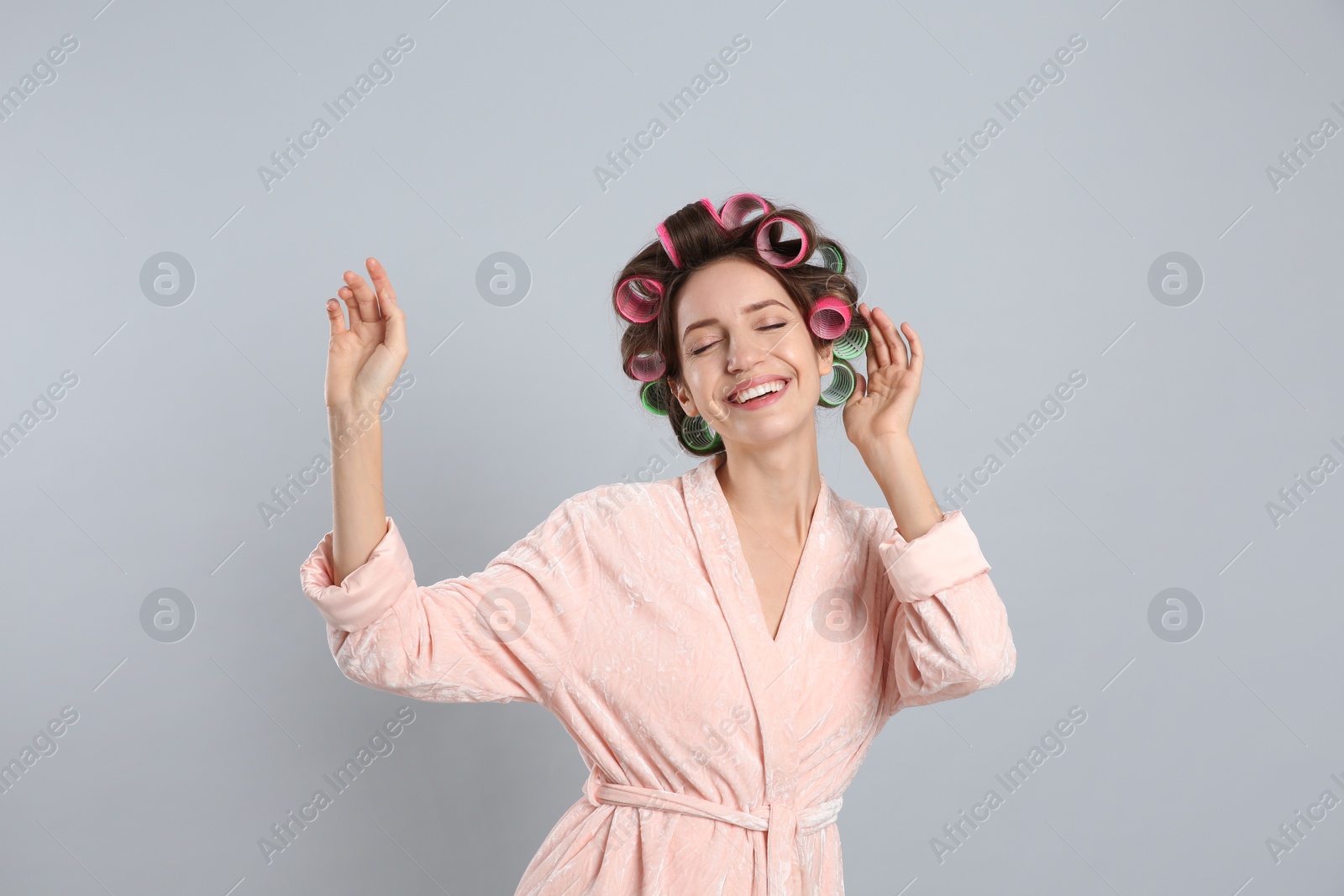 Photo of Happy young woman in bathrobe with hair curlers dancing on light grey background