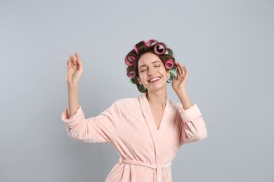 Happy young woman in bathrobe with hair curlers dancing on light grey background