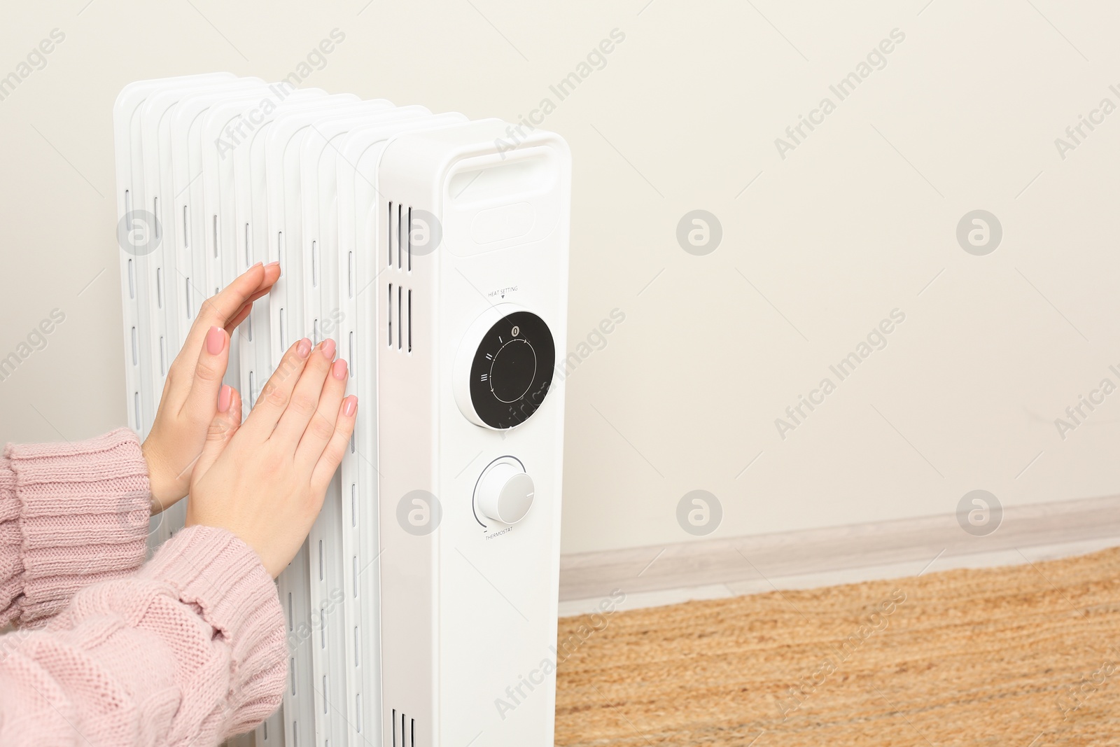 Photo of Young woman warming hands near modern electric heater against beige background indoors, closeup